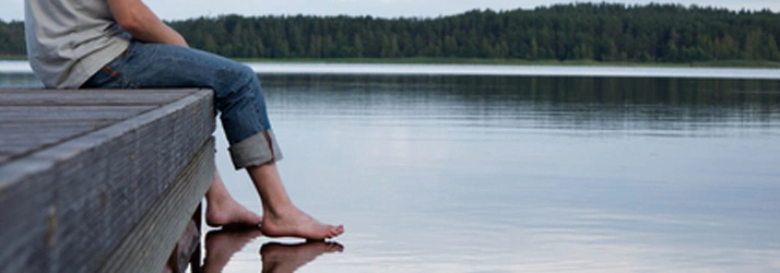 Person sitting on a wooden dock with bare feet touching the water, surrounded by a lake and trees.